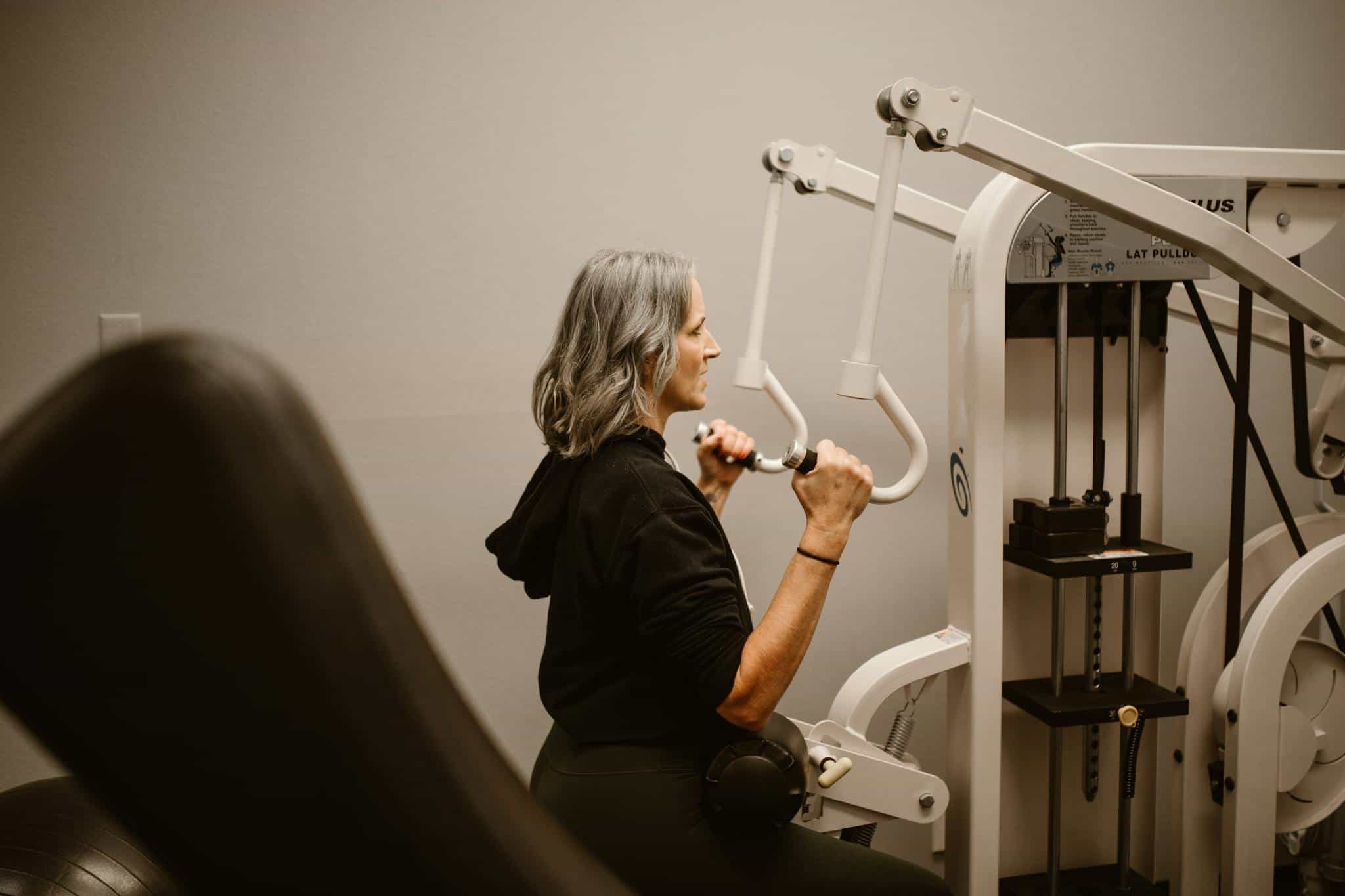 A middle-aged woman with gray hair using a lat pulldown machine for fitness indoors.