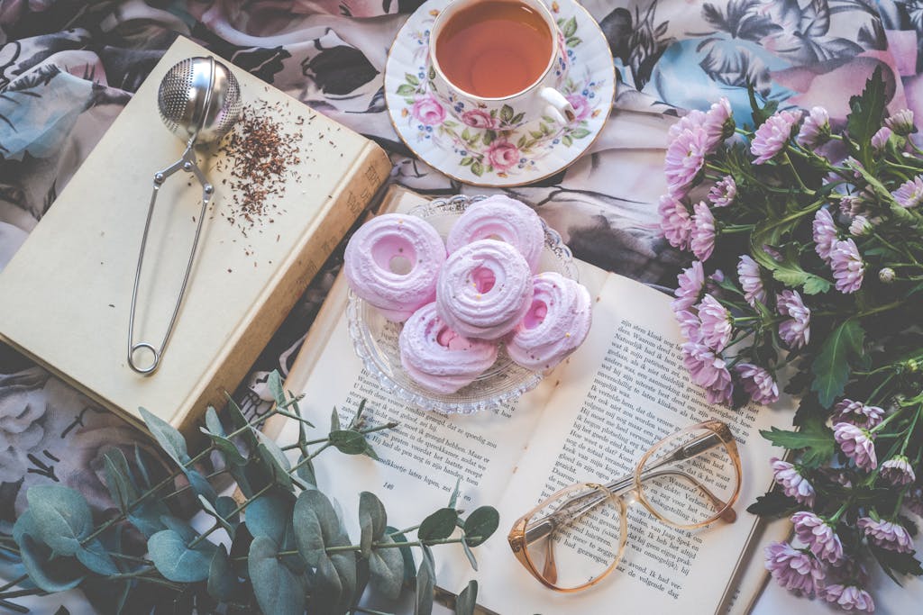 Delicate flatlay with pink pastries, tea, flowers, book, and glasses for a romantic setting.